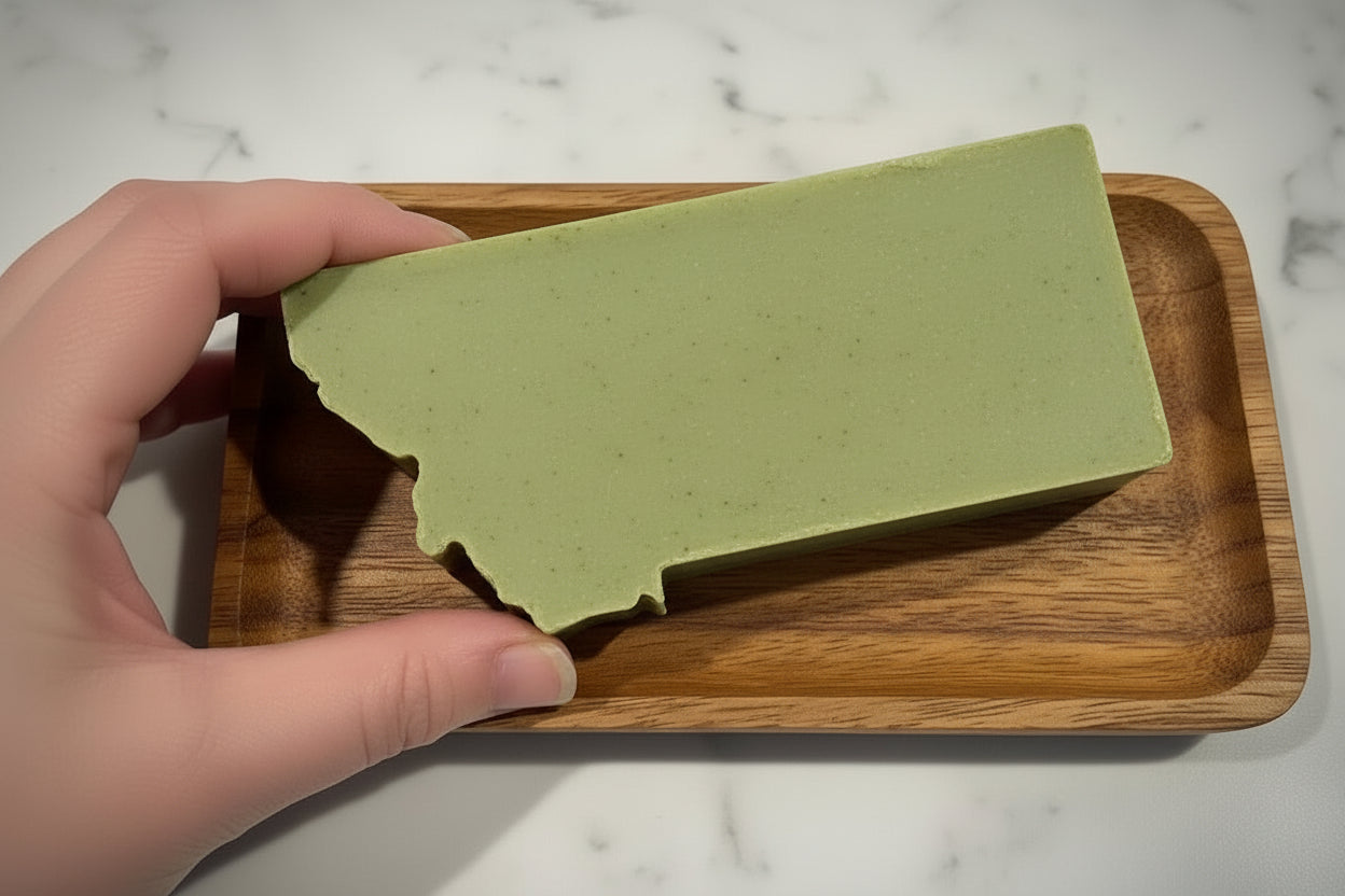 Bar of soap in the shape of the state of Montana wrapped with green  held by a hand on a marble surface