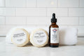 Shampoo bar, conditioner bar, and hair/beard oil bottle on a  white surface in front of a white towel