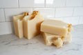 Four bars of handmade soap with a creamy off-white color and small dried botanical inclusions, arranged on a white surface near a window.