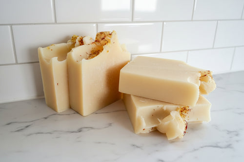 Four bars of handmade soap with a creamy off-white color and small dried botanical inclusions, arranged on a white surface near a window.